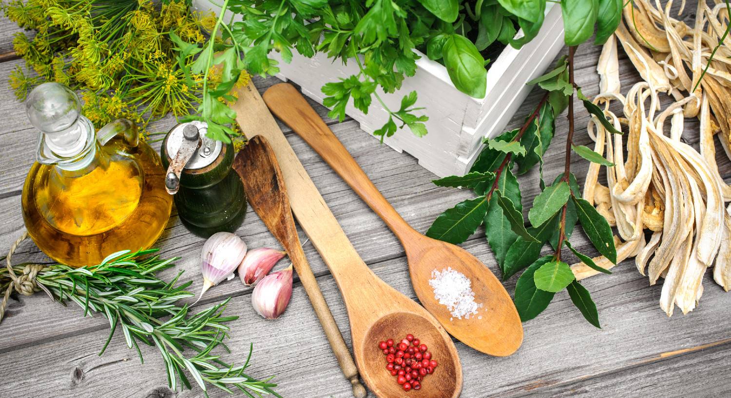 Wooden spoons on a wood table with green herbs and a pepper grinder. 